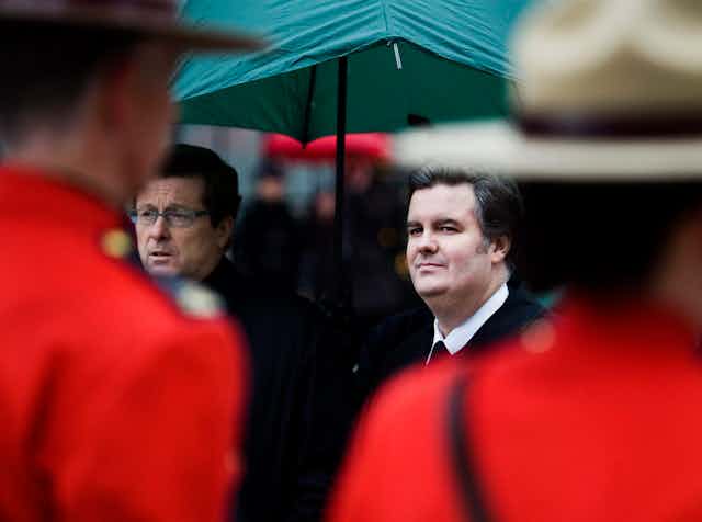 Two men stand under a green umbrella, one frowning and one grinning, as two red-coated Mounties stand in front of them.