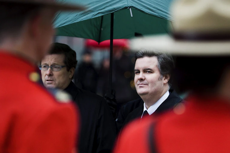 Two men stand under a green umbrella, one frowning and one grinning, as two red-coated Mounties stand in front of them.