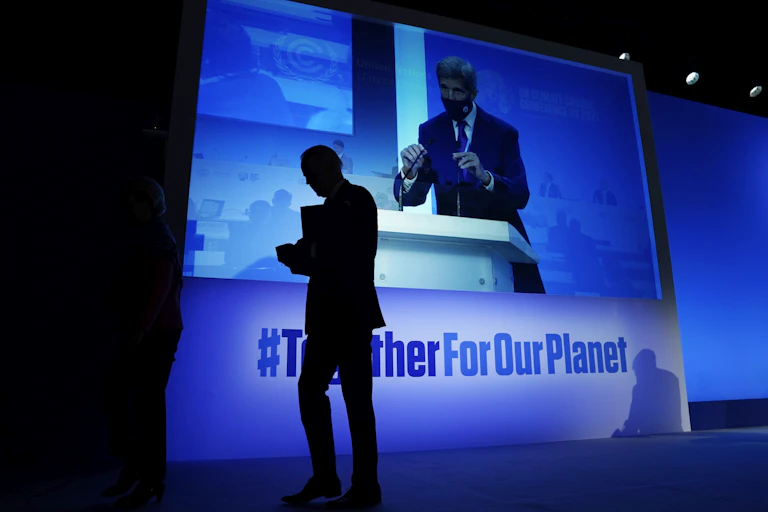 Joe Biden walks past a large screen where John Kerry is speaking to an audience.