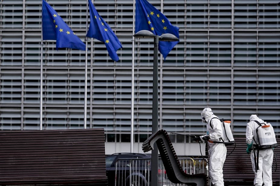 Personnes en tenues antibactériologiques désinfectants des bancs devant un grand bâtiment gris et des drapeaux de l'UE. 