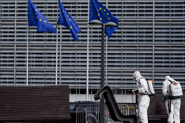 Personnes en tenues antibactériologiques désinfectants des bancs devant un grand bâtiment gris et des drapeaux de l'UE.