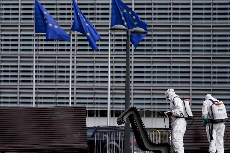 Personnes en tenues antibactériologiques désinfectants des bancs devant un grand bâtiment gris et des drapeaux de l'UE.