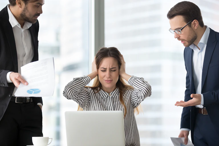 A white woman sits behind a laptop with her hands over her ears, eyes closed. Two men stand on either side of her, demanding her attention.