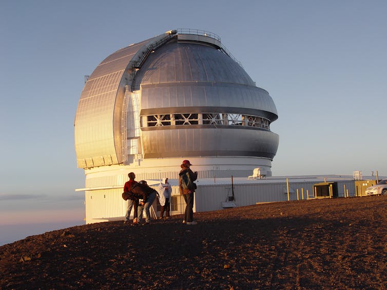 Researchers stand next to a large domed observatory on barren mountaintop at sunset.