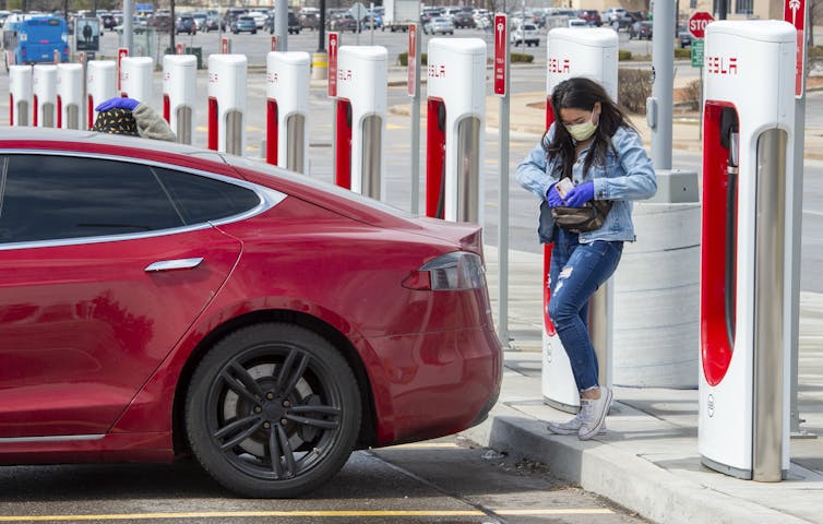 A woman stands behind her red tesla electric car and reaches into her hip pouch as she prepares to plug it into one of the many charging spots.