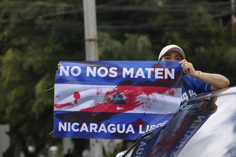 A protester leans out of a car holding a anti-Ortega banner reading 'do not kill us.'
