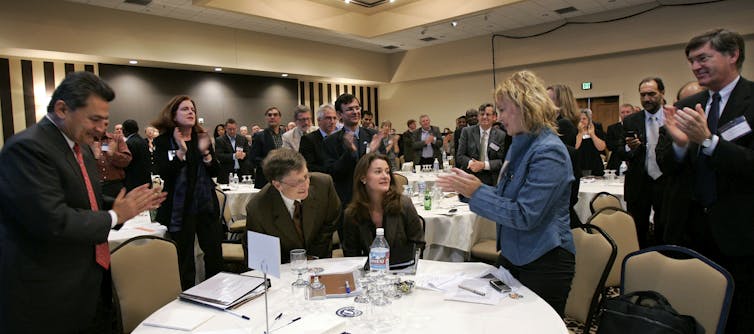 A roomful of people in suits applaud Bill and Melinda Gates