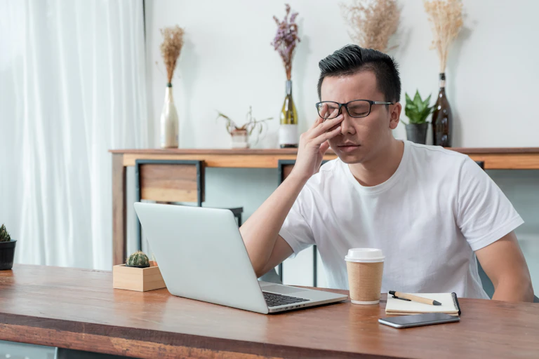 An Asian man sits behind his laptop with his hand over his face looking exhausted