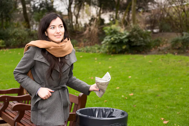 A white woman wearing a fall jacket and scarf throws a crumpled up newspaper in the trash.