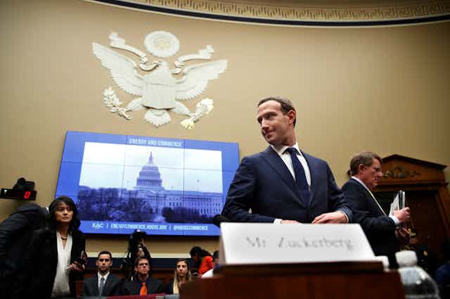 A man in a business suit stands behind a name card that reads Mr. Zuckerberg as people stand or sit in the background and a video screen on the wall displays an image of the U.S. Capitol building