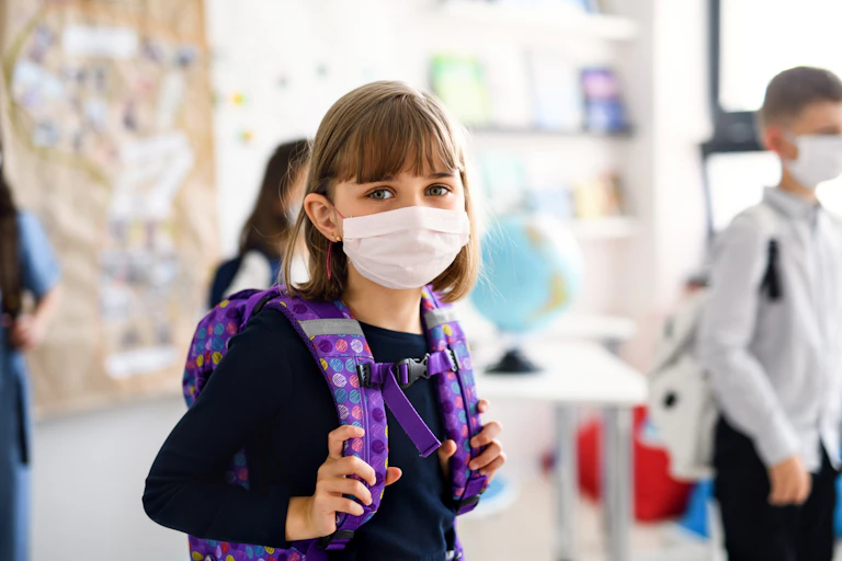 Eight year old girl in a mask and backpack looks at the camera.