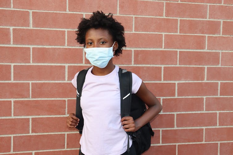 Afrian Australian child in a light pink top wearing a mask stands in front of a brick wall.