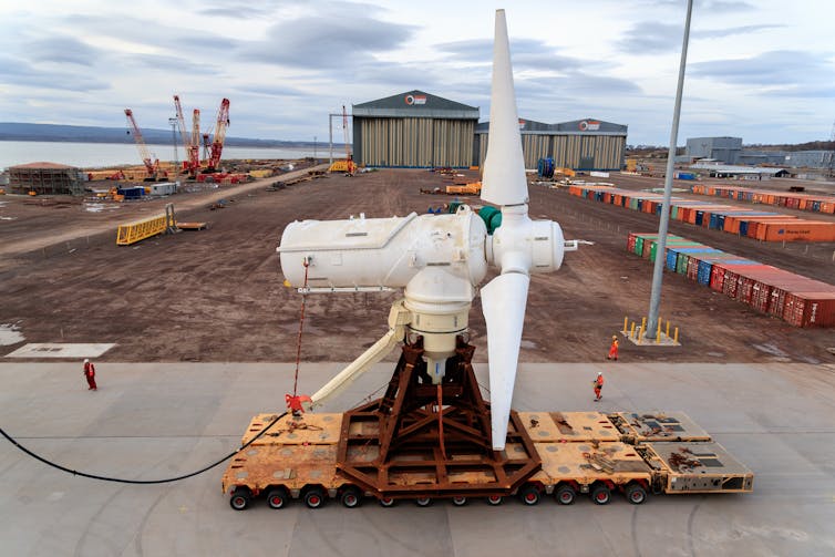 A large white turbine on a metal pallet surrounded by shipping containers.