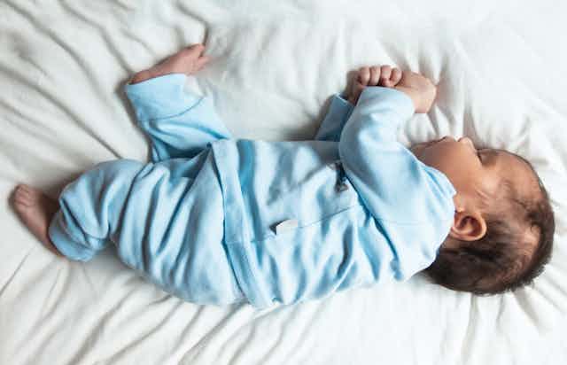 A small baby sleeps on their side wearing blue pajamas on a white sheet