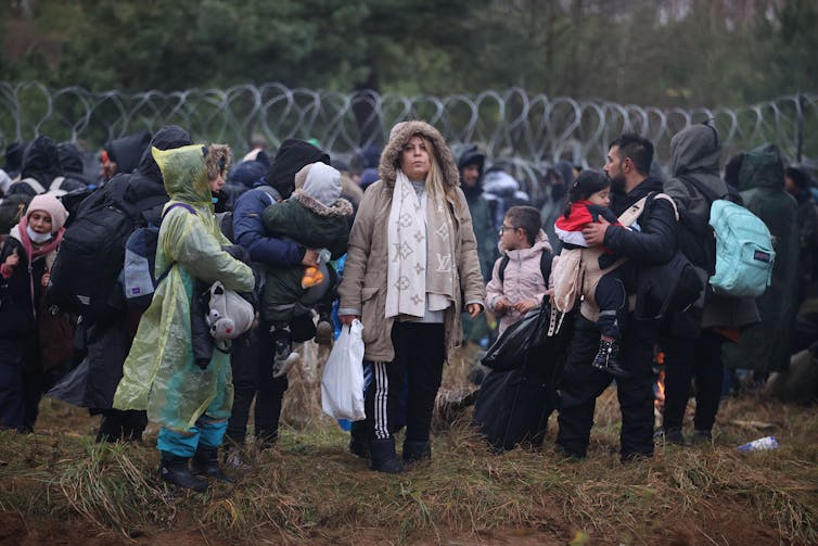 Men, Women And Children In Winter Clothes Stand In Front Of The Barbed Wire Border Between Belarus And Poland.
