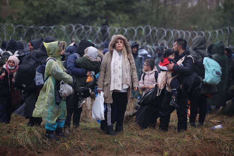 Men, women and children in winter clothing standing in front of the barbed-wire border between Belarus and Poland.