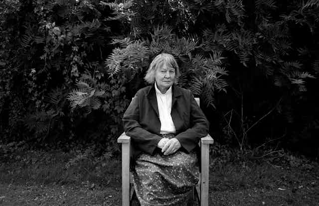 A black and white photo of Iris Murdoch seated in a chair in her garden, looking towards the camera