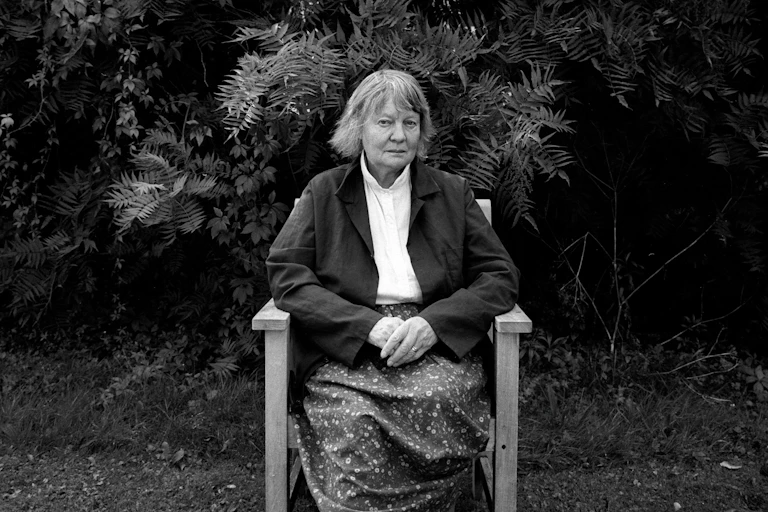 A black and white photo of Iris Murdoch seated in a chair in her garden, looking towards the camera