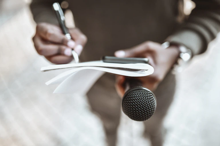 A man holding a mobile phone and microphone and writing in a notebook.