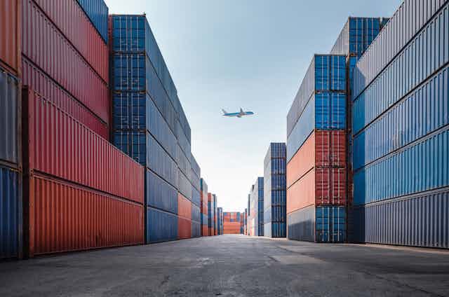Unmarked freight containers stacked high at a shipping port with an airplane in the background