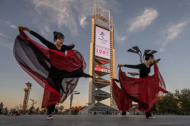 Women in red and black outfits rehearsing a dance in front of an Olympic sign in Beijing, China