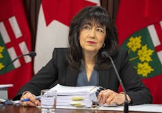 A woman with dark hair sits at a desk behind a microphone with a large binder in front of her and flags behind her.