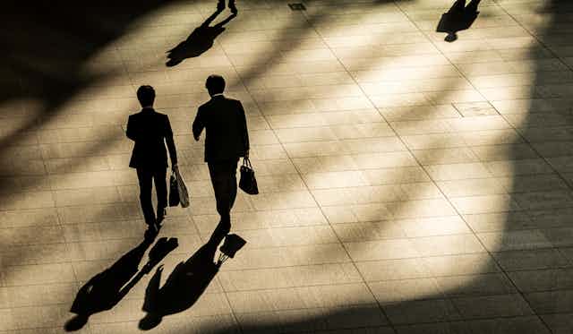 Two men walk carrying briefcases in the shadows of buildings.