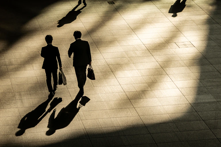 Two men walk carrying briefcases in the shadows of buildings.