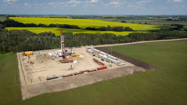 An aerial view of farmland and some woods, with a large square section carved out and containing an oil rig and several shipping containers of equipment. 