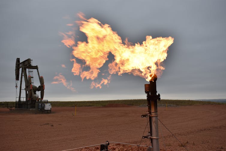 A flare burns natural gas at an oil well with a drilling rig in the background.