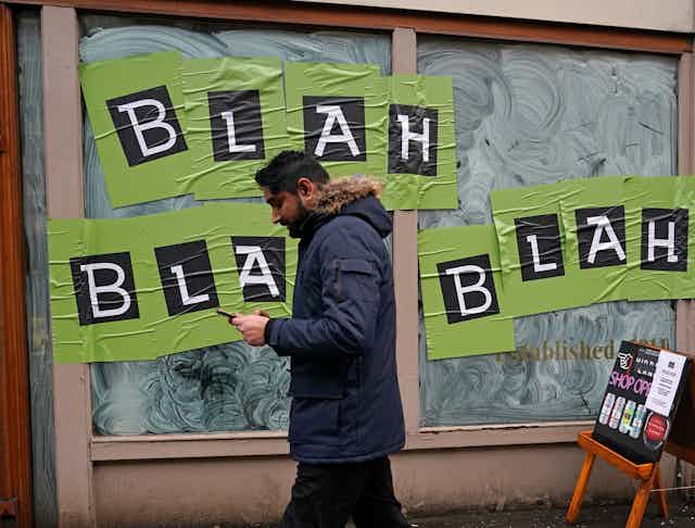 A man looks at his smartphone while walking past a window with a sign reading: Blah, blah, blah