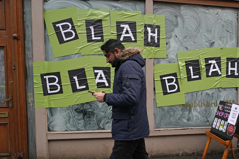 A man looks at his smartphone while walking past a window with a sign reading: Blah, blah, blah