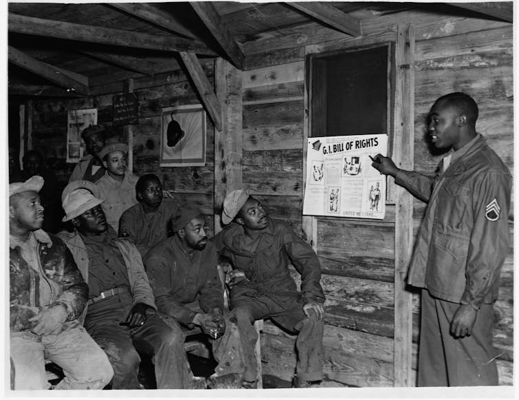 A Black man points to a sign about the G.I. Bill of Rights while speaking to a group of African American men.
