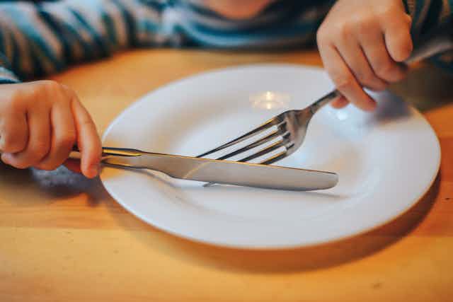 Child's hands hold a fork and knife across an empty plate.