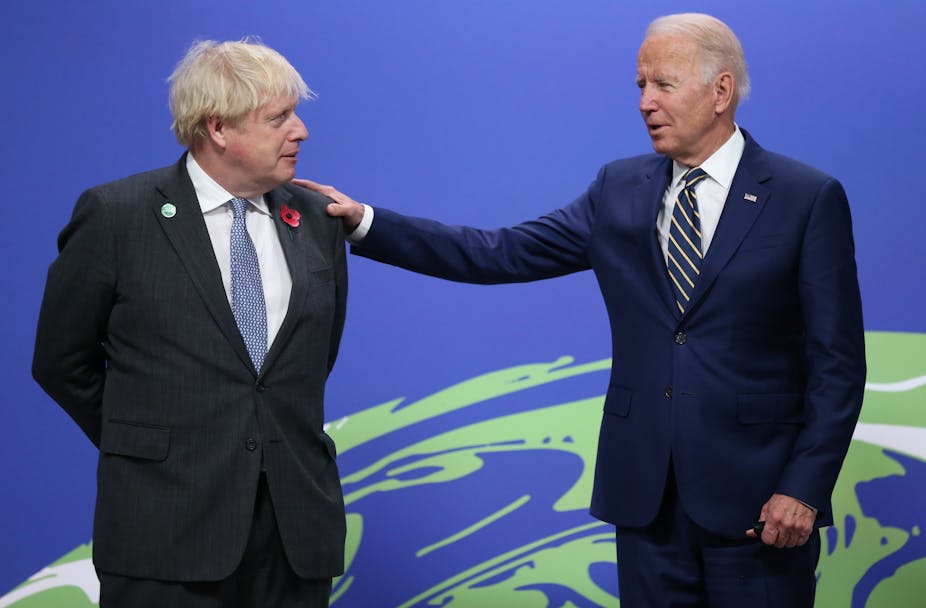 Joe Biden rests his right hand on Boris Johnson's left shoulder as the two leaders greet each other in front of a blue and green background.