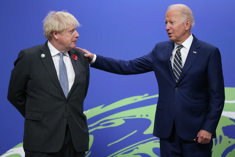 Joe Biden rests his right hand on Boris Johnson's left shoulder as the two leaders greet each other in front of a blue and green background.