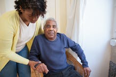 A woman assists an elderly man sitting in a chair by a window.