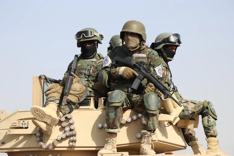 Troops in camo with guns sit atop a tank