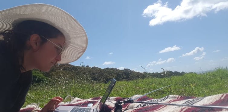 A person wearing a hat lies on a blanket in a vast field on a sunny day with a laptop and antenna.