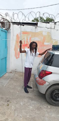 A person holds an antenna aloft with one hand on a laptop on the roof of a car.