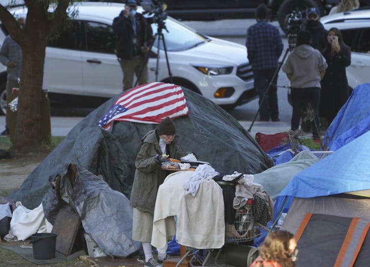 a woman eats at her tent adorned with an American flag at the Echo Park homeless encampment at Echo Park Lake in Los Angeles. California