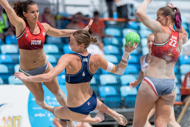 A woman wearing a red bikini top with grey bottom flies through the air as she tries to block a shot.