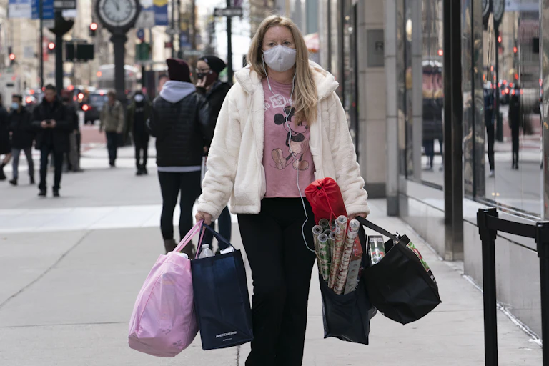 a woman wearing a mask carries four shopping bags stuff with stuff on the sidewalk in New York.