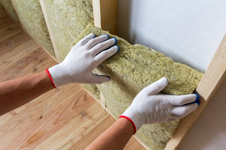 Gloved hands fitting yellow foam insulation into a wall cavity.
