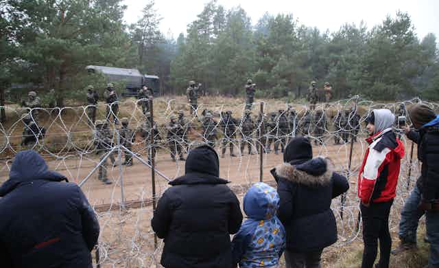 Adults and children in coats look through barbed wire fence at line of soldiers