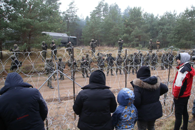 Adults and children in coats look through barbed wire fence at line of soldiers