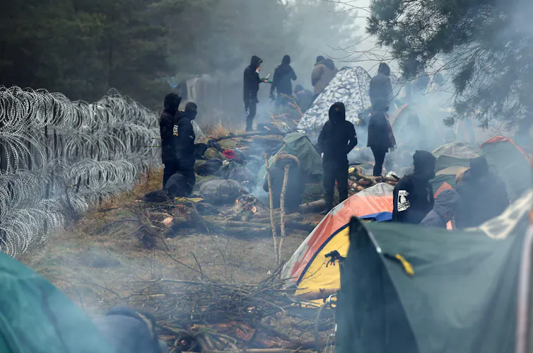 Men in black hoodies and masks next to tents and piles of firewood, alongside a barbed wire fence.