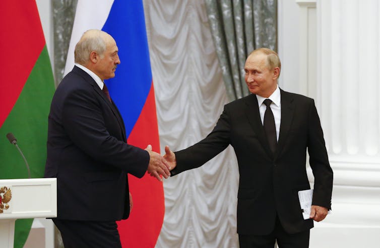 Alexander Lukashenko shakes hands with Vladimir Putin in front of Russian and Ukrainian flags.