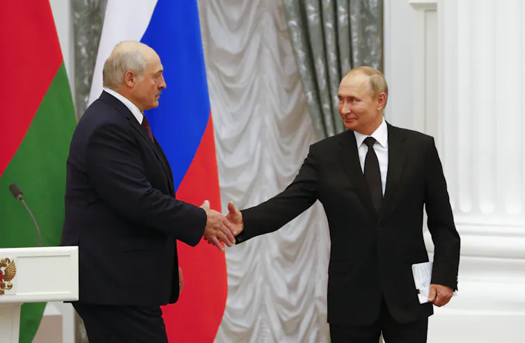 Alexander Lukashenko shakes hands with Vladimir Putin in front of Russian and Ukrainian flags.