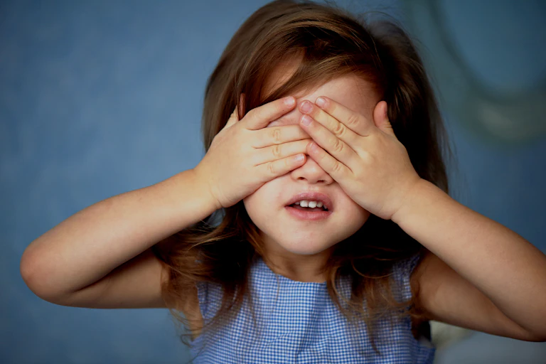 Niña con vestido azul se tapa los ojos.